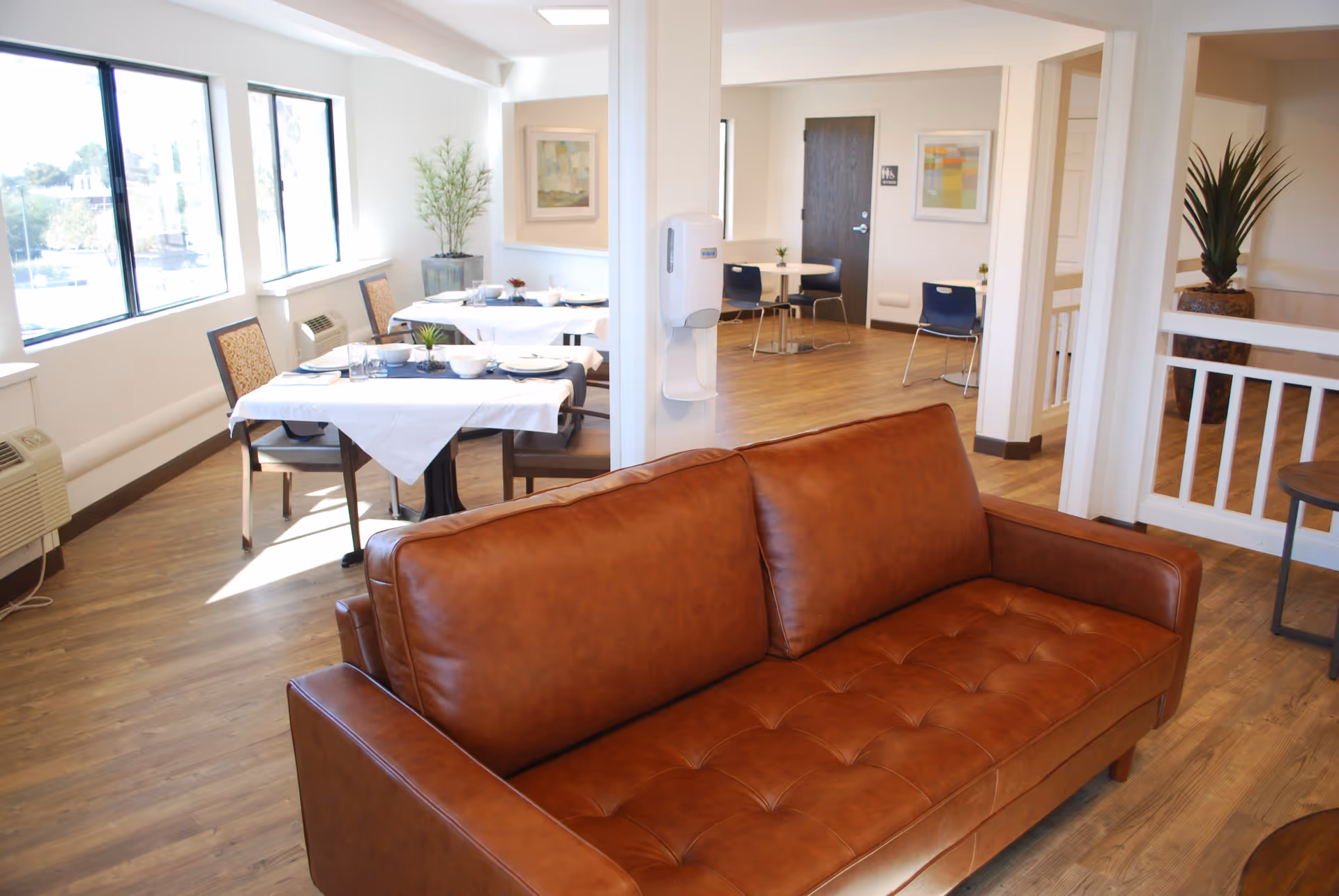 Interior view of a senior living facility common area with a brown leather sofa in the foreground and dining tables set with white tablecloths and place settings near large windows. The room has wooden flooring, potted plants, and modern decor with bright natural light.