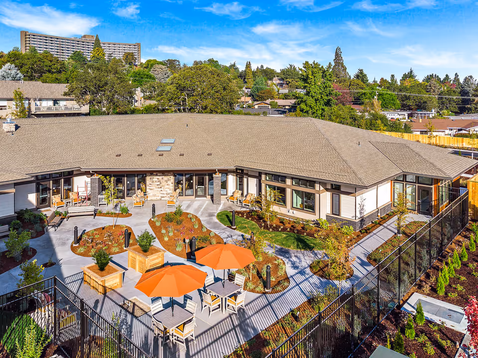 Landscaped outdoor courtyard with patio tables and orange umbrellas surrounded by a single-story memory care building.