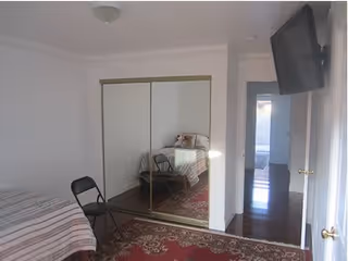 A small bedroom with a bed covered in a striped blanket, a black folding chair, a large mirrored closet door, a red patterned rug on the floor, and a wall-mounted TV. The room has white walls and a doorway leading to another area with wooden flooring.