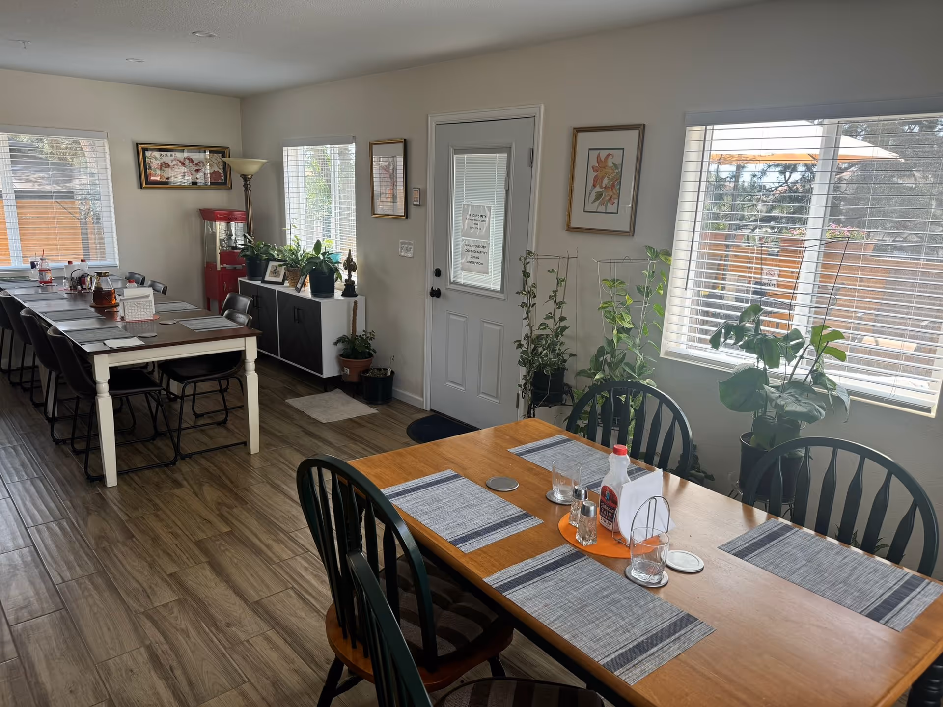 A bright dining room in Lotus Assisted Living with two wooden tables set with placemats, glasses, and condiments. The room has large windows with blinds letting in natural light, several potted plants near the windows and door, framed artwork on the walls, and a popcorn machine in the corner. The floor is wood-patterned and there are multiple chairs around the tables.