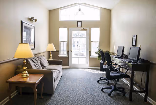 A bright room with a beige sofa and a wooden side table with a lamp on the left side, and a long desk with two computer workstations and office chairs on the right side. The room has a large window and a glass door letting in natural light, with a plant near the door.