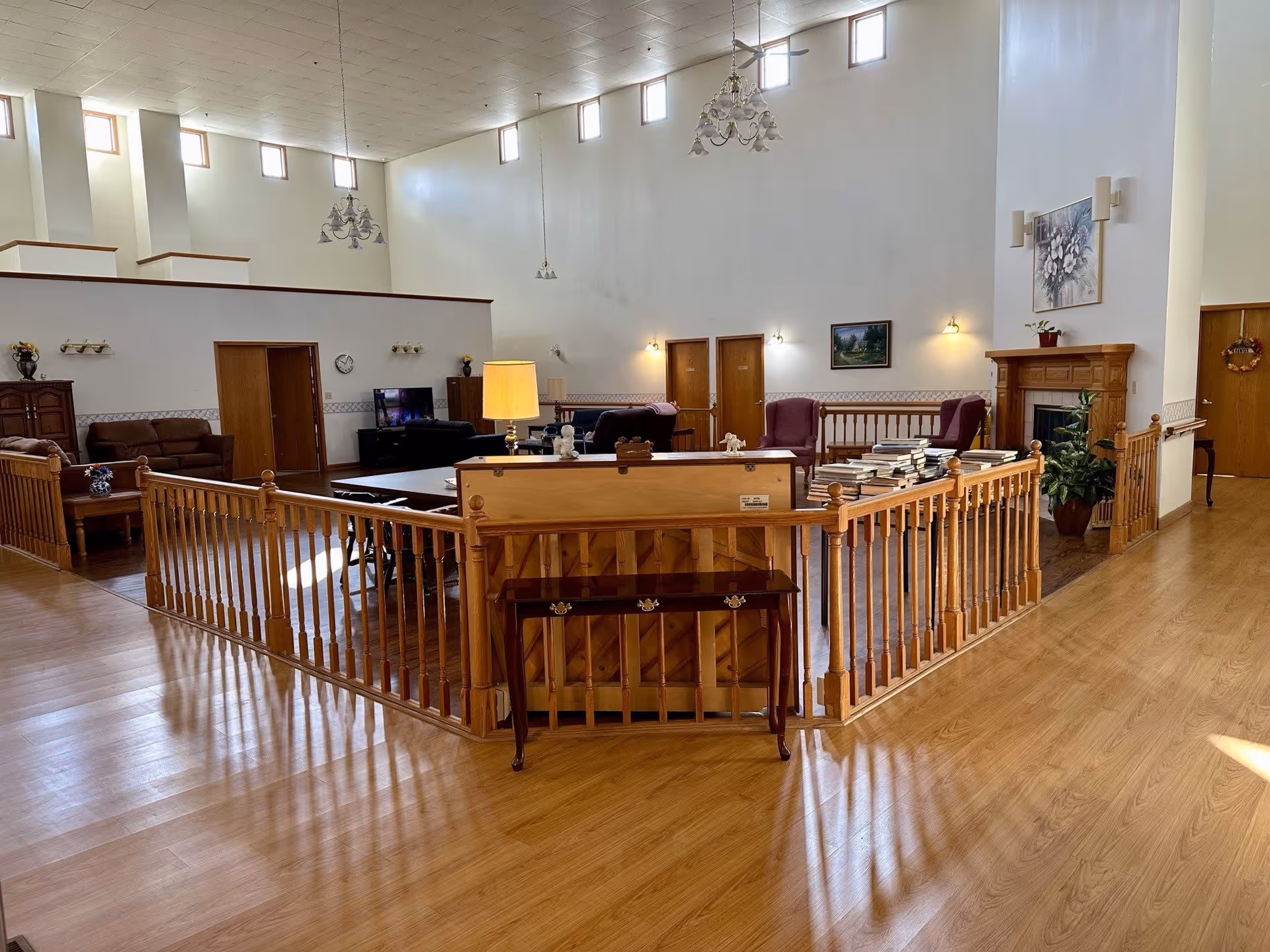 A spacious senior living facility common area with high ceilings and multiple windows near the ceiling allowing natural light. The room features wooden flooring, a wooden railing enclosing a seating area with sofas, armchairs, a piano, a table lamp, and a fireplace with a painting above it. There are doors along the walls and decorative wall sconces providing additional lighting.