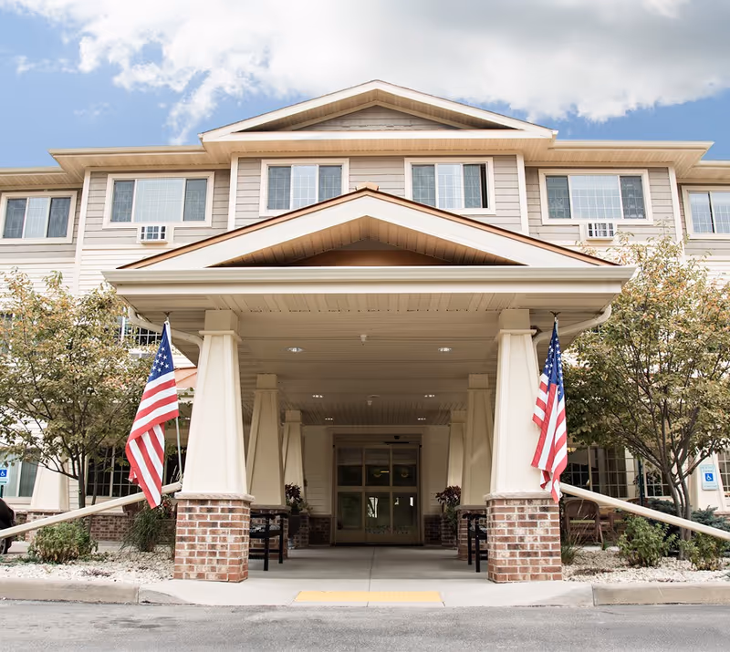 Covered main entrance of a multi-story assisted living building with a portico, two American flags, and glass double doors.