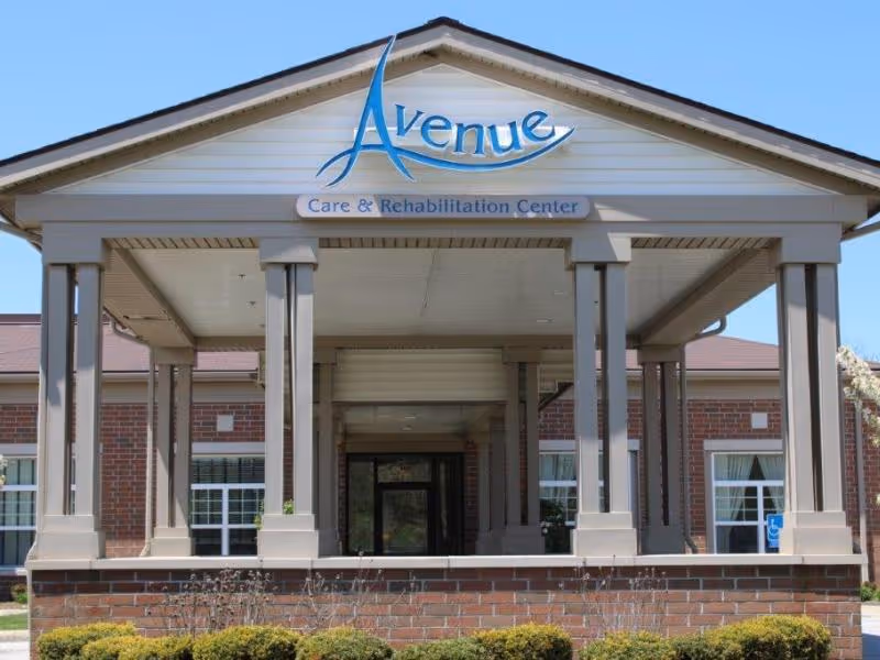Front exterior view of Avenue Care & Rehabilitation Center showing a covered entrance with columns, brick walls, windows with white curtains, and a clear blue sky.
