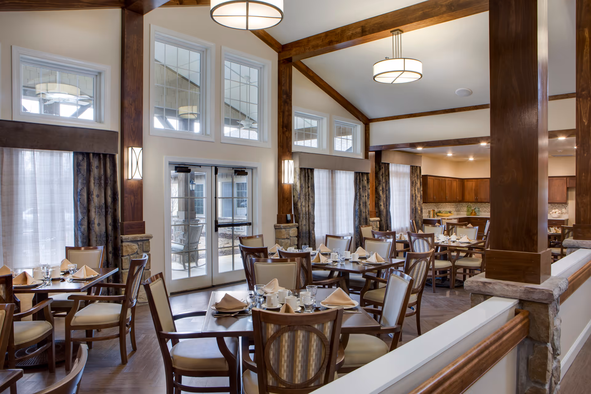 A spacious dining room with multiple wooden tables and chairs arranged neatly. Each table is set with beige napkins, white cups, glasses, and silverware. The room features large windows with curtains, wooden beams on the ceiling, and modern hanging light fixtures. There is a door leading outside and a kitchen area visible in the background.