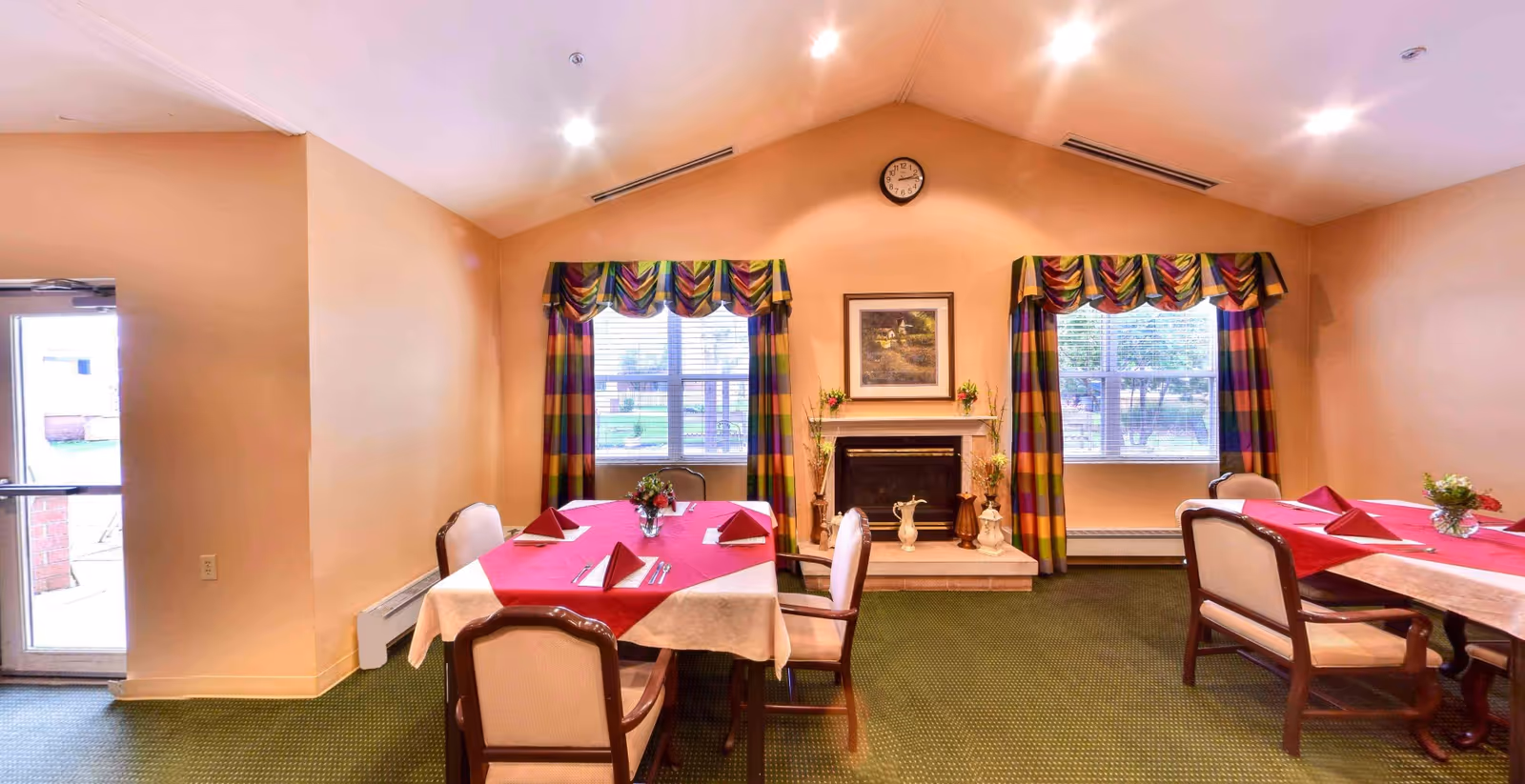 A cozy dining room with two tables covered in white and red tablecloths, each set with folded red napkins, silverware, and small flower arrangements. The room has green carpet, peach-colored walls, two windows with colorful plaid curtains, a fireplace with decorative items on the mantel, and a clock above it. There is a door to the left leading outside.