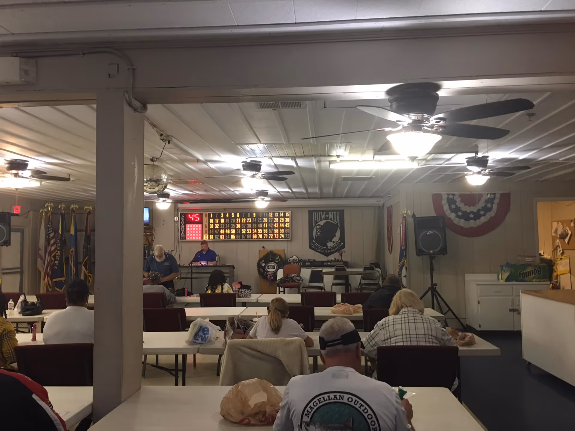A group of people sitting at tables in a room playing bingo. The room has ceiling fans with lights, a large electronic bingo board on the wall, and several flags displayed on the left side. There is a person standing near a microphone at the front of the room, and various items like bags and food are on the tables.