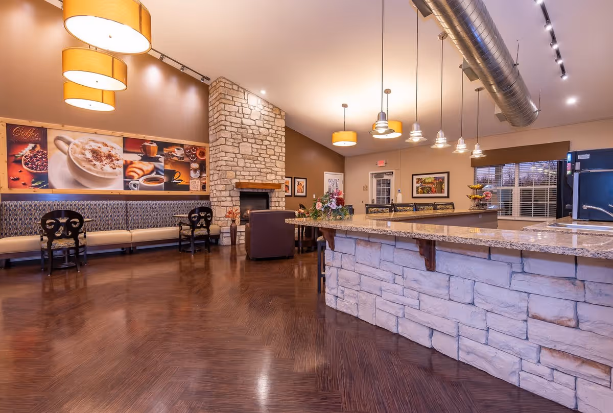 Interior of a senior living facility common area with a stone fireplace, a long cushioned bench with patterned upholstery, several chairs, a large stone and granite countertop bar with hanging pendant lights, and wall art featuring coffee-themed images.