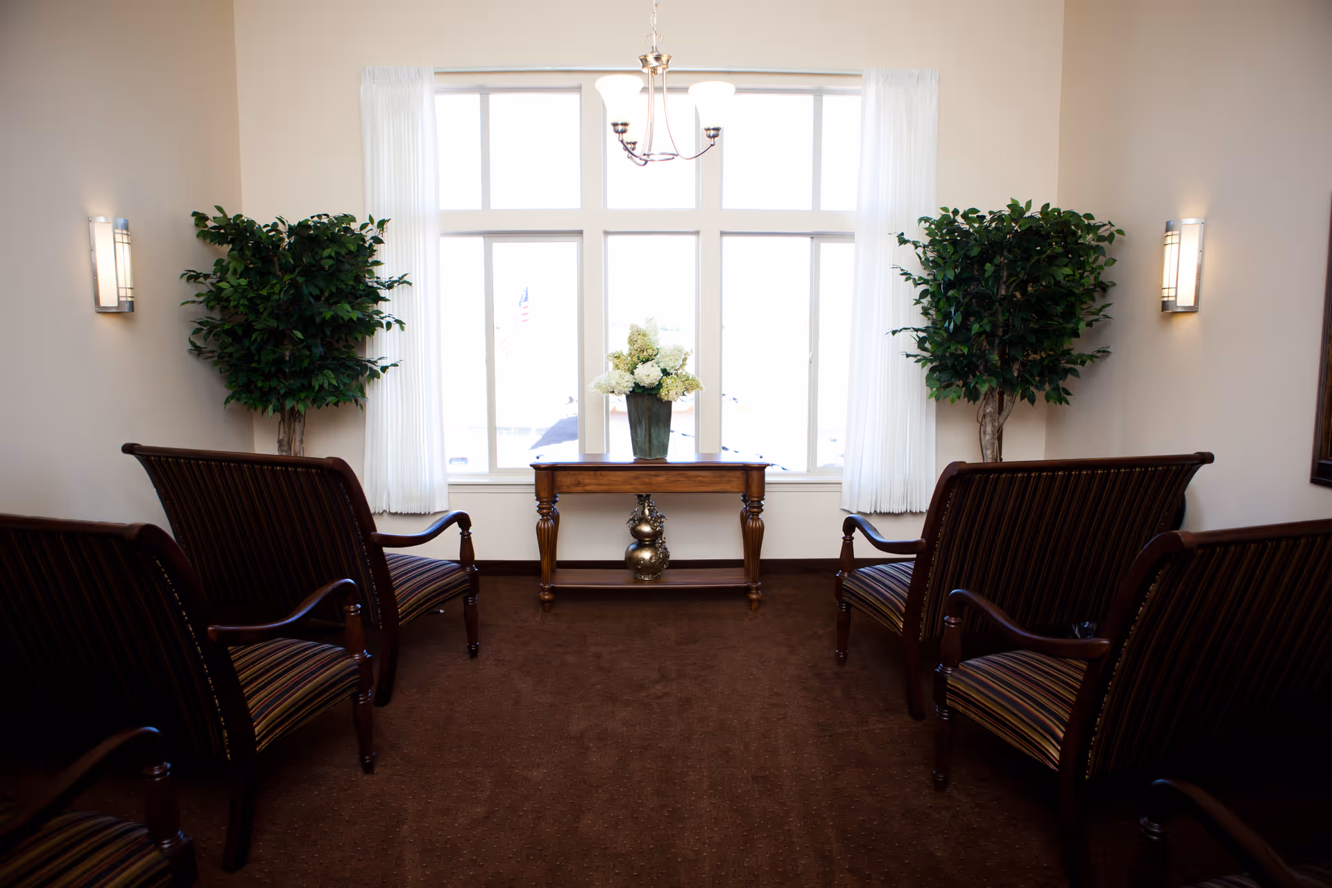 A small, well-lit sitting area with four wooden armchairs upholstered in striped fabric arranged facing each other. Between two large windows with white curtains is a wooden table with a vase of white flowers. Two tall green plants are placed on either side of the windows, and wall sconces provide additional lighting.