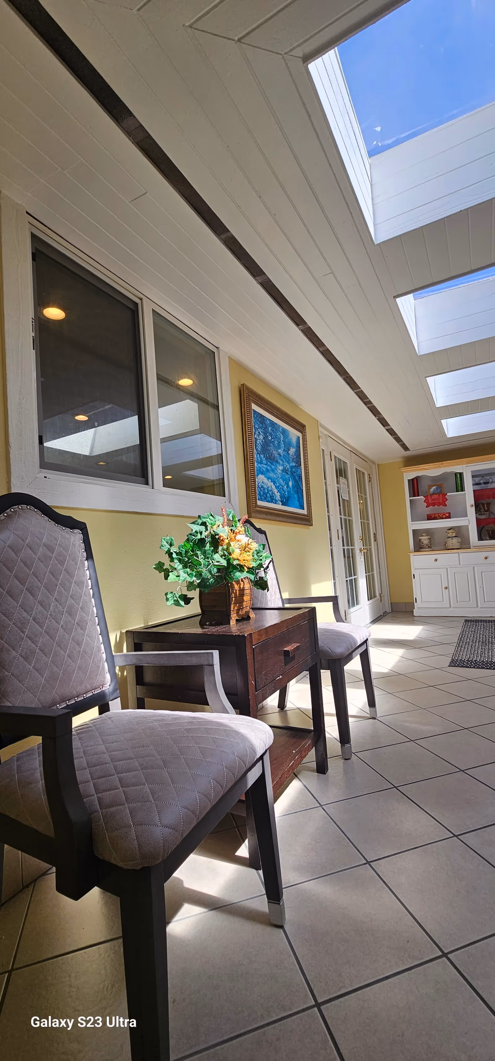 A bright indoor hallway with two cushioned chairs and a small wooden table with a flower arrangement. The walls are painted yellow, and there is a framed blue artwork hanging on the wall. The ceiling features multiple skylights allowing natural light to fill the space. At the end of the hallway, there is a white cabinet with shelves and glass doors.