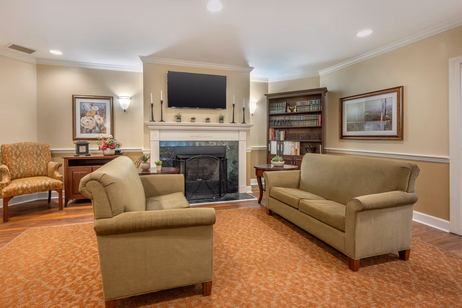 A cozy living room with two beige sofas facing each other on an orange patterned carpet, a fireplace with a black screen and a flat-screen TV mounted above it, a wooden bookshelf filled with books, a side table with a plant, a patterned armchair, and framed artwork on the beige walls.
