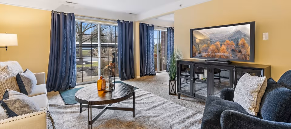 Well-lit living room with a sofa and armchair, round coffee table, TV on a console, and large windows with dark curtains.
