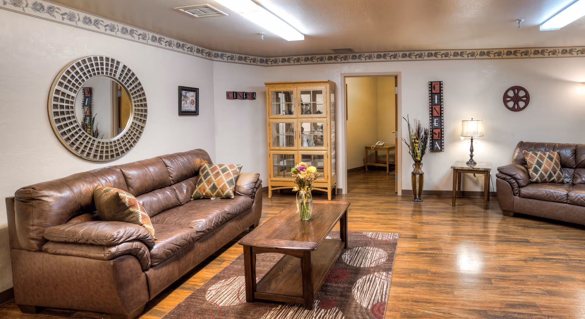 A cozy living room with two brown leather sofas adorned with patterned cushions, a wooden coffee table with a vase of flowers, a decorative round mirror on the wall, a wooden display cabinet, a floor vase with flowers, a side table with a lamp, and wall decorations including a 'CINEMA' sign and a film reel. The room has wooden flooring and soft lighting.