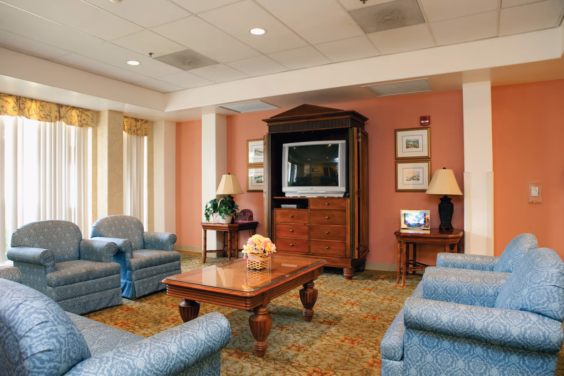 Bright senior-living common room with upholstered armchairs arranged around a wooden coffee table and a TV armoire.