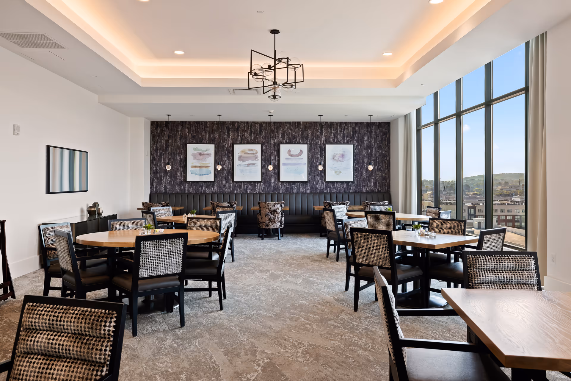 Bright dining room with multiple tables and chairs, large windows and framed artwork on the far wall.