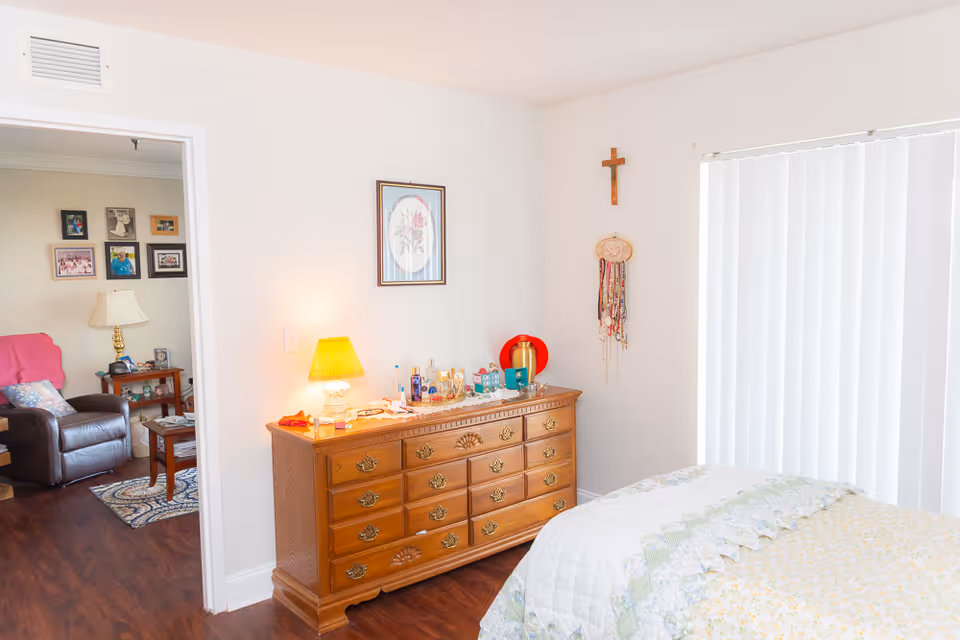 A bright bedroom with a wooden dresser topped with a lamp, perfumes, and other small items. A bed with a patterned quilt is partially visible on the right. A cross and hanging necklaces are on the wall above the dresser. The room has white walls and vertical blinds covering a window. An open doorway leads to a living room area with a leather recliner, side table with a lamp, and framed photos on the wall.