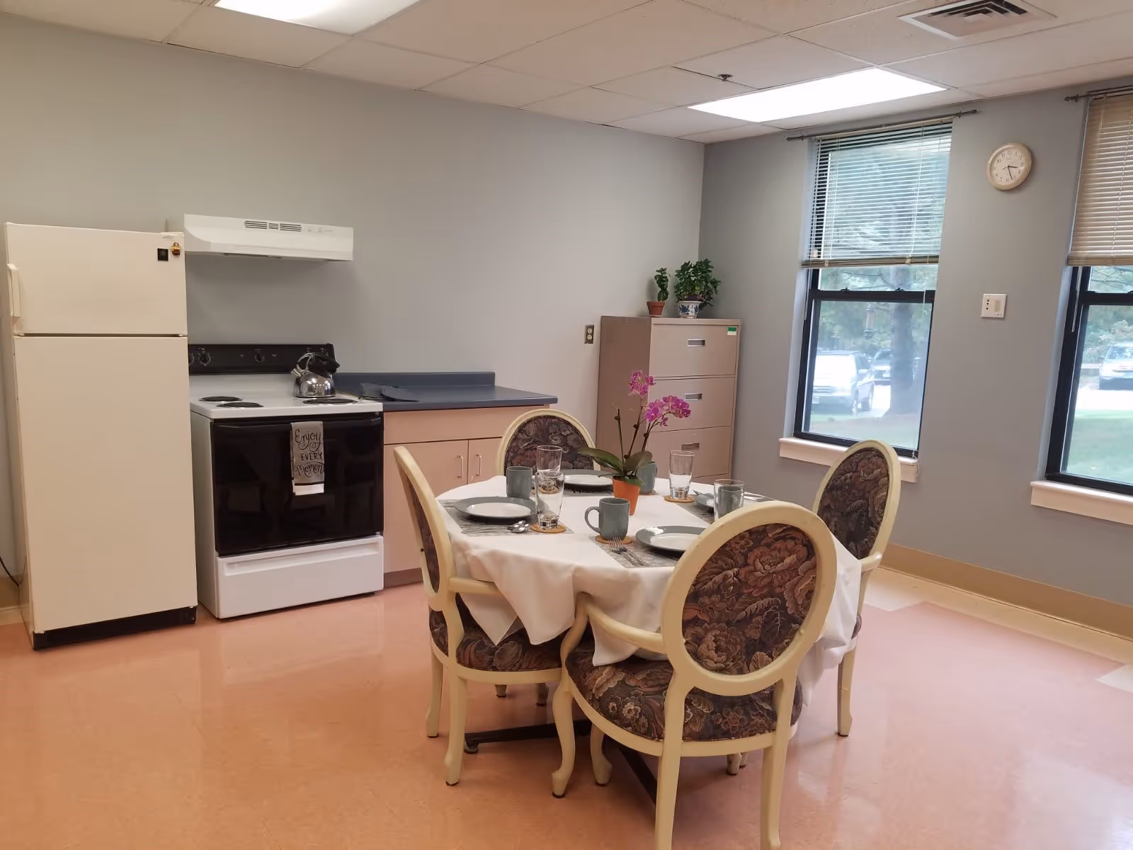 A small kitchen and dining area with a white refrigerator, black and white stove with a kettle on it, and a countertop with cabinets. In the center, there is a round table covered with a white tablecloth, set with plates, glasses, and mugs, and surrounded by four upholstered chairs with floral patterns. Two windows with blinds let in natural light, and a clock is mounted on the wall above a filing cabinet with potted plants on top.