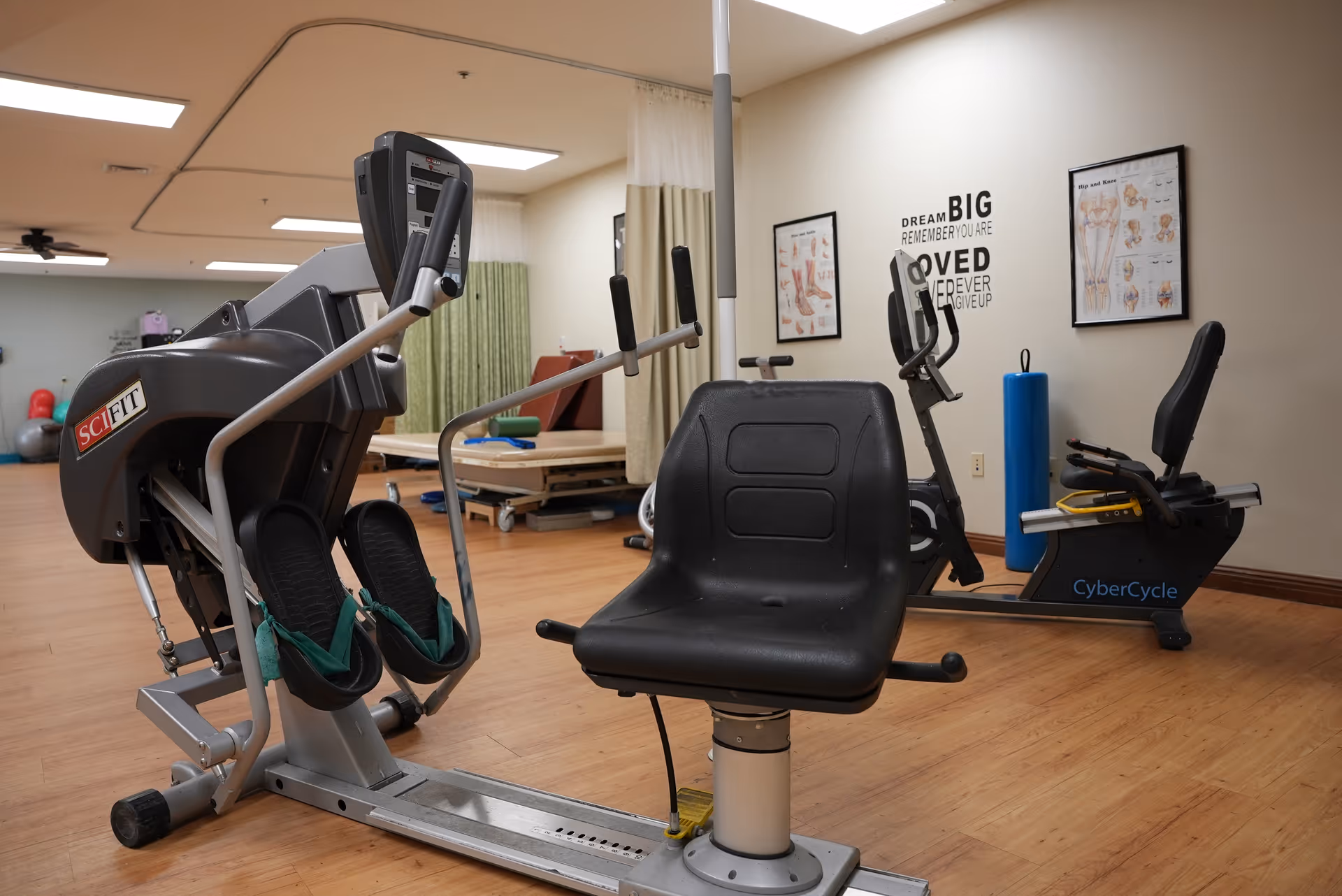 A fitness room with exercise equipment including a SciFit machine and a CyberCycle stationary bike. The room has wooden flooring, anatomical posters on the wall, and a motivational quote that reads 'Dream Big Remember You Are Loved Never Give Up'.
