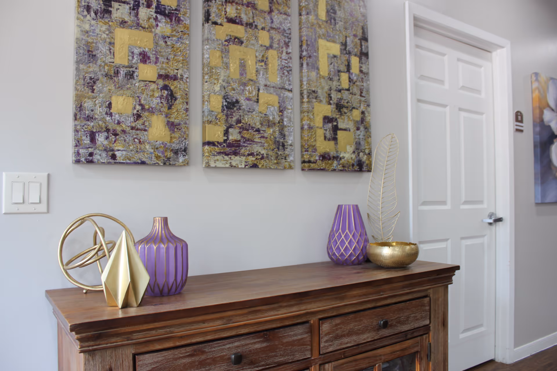 A wooden sideboard with decorative items including two purple vases, a gold geometric sculpture, a gold leaf ornament in a small gold bowl, and a gold circular sculpture. Above the sideboard are three abstract paintings with purple and gold patterns. To the right is a white door with a silver handle.