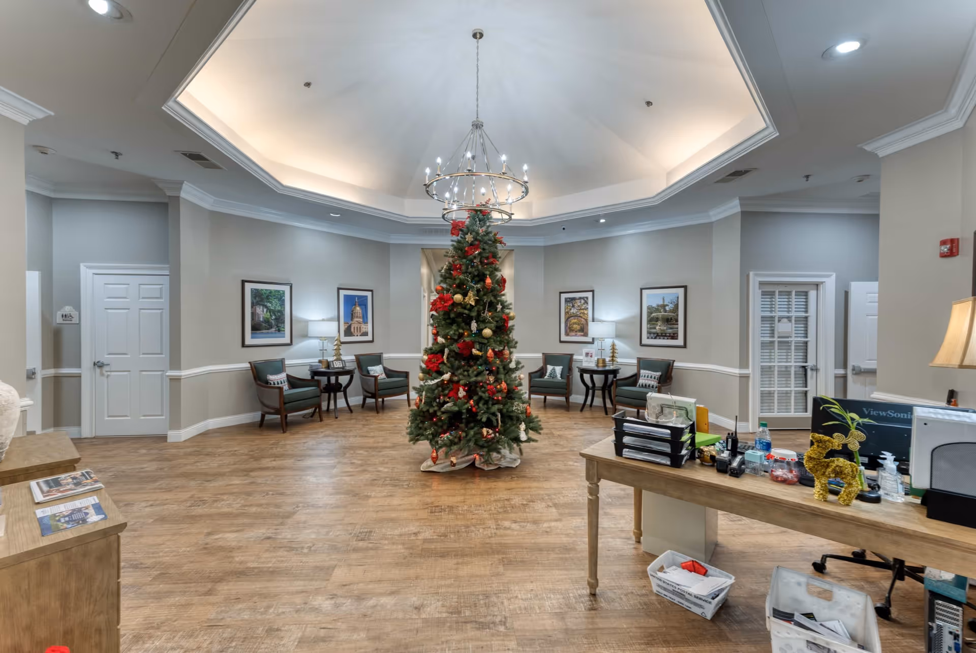 A spacious, well-lit common area decorated for Christmas with a large Christmas tree in the center. The room features wooden flooring, several green cushioned chairs with small tables and lamps along the walls, framed pictures, and a chandelier hanging from a recessed ceiling. A desk with office supplies and holiday decorations is visible on the right side.