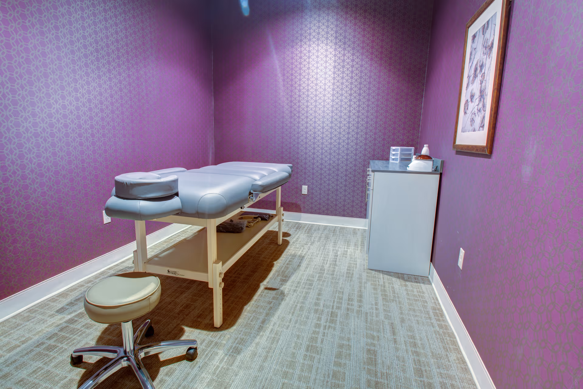 A small room with purple patterned walls featuring a massage table with a face cradle and a rolling stool. There is a cabinet with some items on top and a framed picture hanging on the wall.