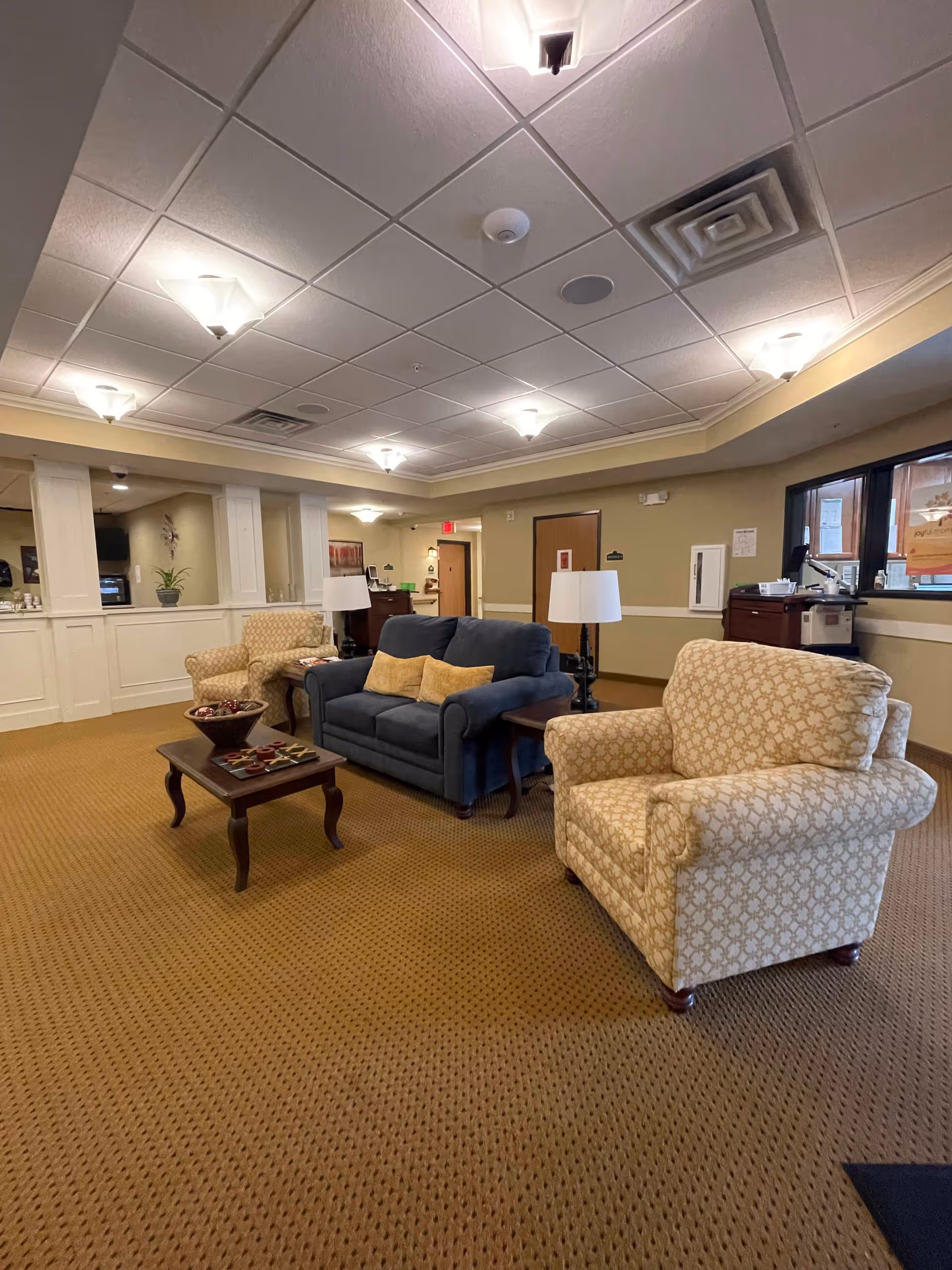 A cozy common area in a senior living facility featuring a blue loveseat with two yellow cushions, two patterned armchairs, a wooden coffee table with decorative items, and two side tables with lamps. The room has beige walls, a carpeted floor, and a ceiling with multiple light fixtures. There is a window on the right side and a hallway leading to other rooms in the background.