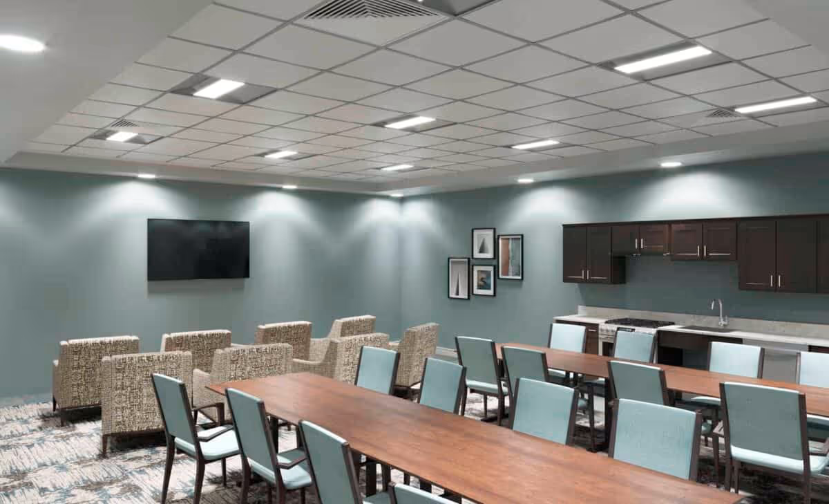 A spacious room with multiple rows of chairs and tables arranged for group seating. The room features a wall-mounted flat-screen TV, a kitchenette area with dark cabinets, a sink, and a stove. The walls are painted light blue, and there are framed pictures on one wall. The ceiling has a grid pattern with recessed lighting.