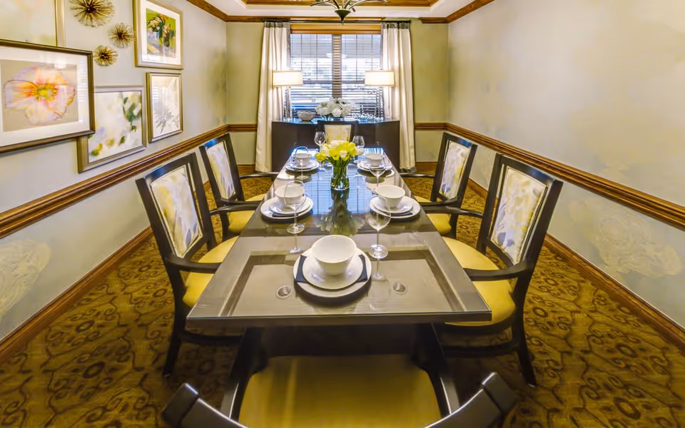 A formal dining room with a long rectangular glass table set for six people. The table has white bowls, plates, wine glasses, and a vase with yellow flowers in the center. Six wooden chairs with yellow cushioned seats and floral-patterned backs surround the table. The room has beige walls with wooden trim, a patterned carpet, framed floral artwork on one wall, and a window with white curtains and two table lamps on a sideboard beneath it.