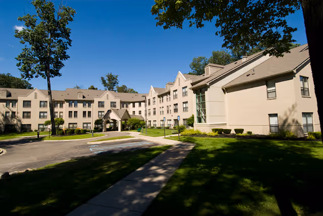 Exterior view of McGivney-Bethune Apartments, a multi-story residential building with beige walls and a sloped roof, surrounded by green lawns, trees, and a paved driveway under a clear blue sky.