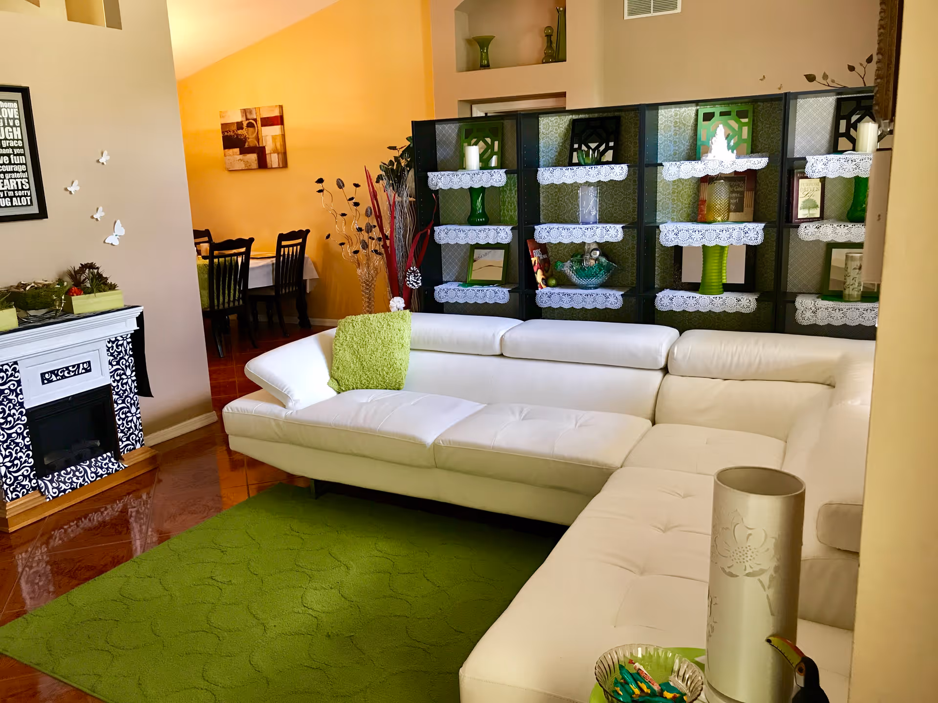 A cozy living room with a large white sectional sofa adorned with a green throw blanket. The room features a green area rug on a polished wooden floor, a decorative black and white fireplace, and a black shelving unit with lace-trimmed shelves displaying various green and white decorative items. In the background, there is a dining area with a table and chairs against a yellow wall.