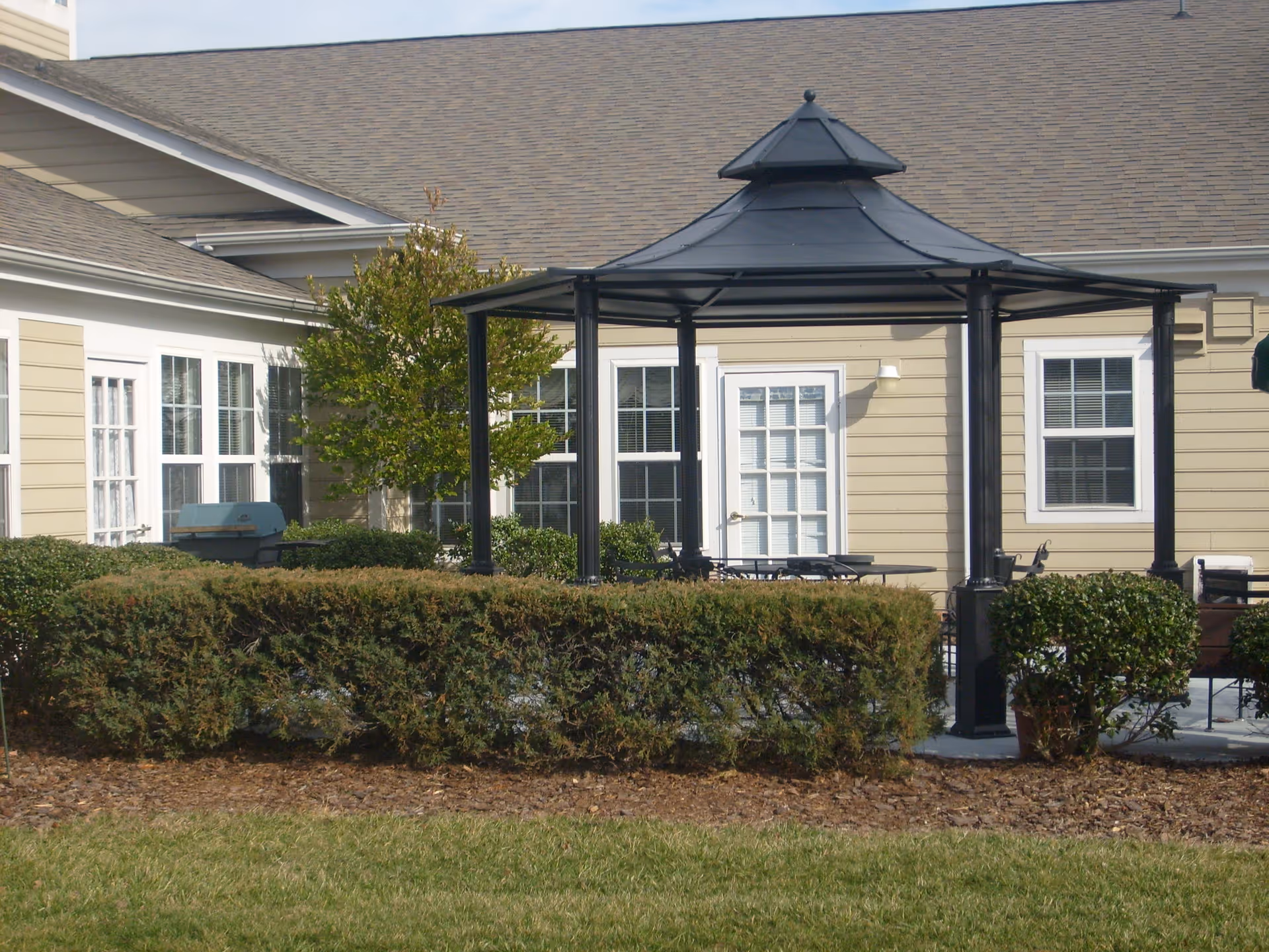 Black metal gazebo on a patio in front of a beige building with windows, shrubs, and a lawn.