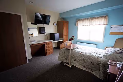 Single bedroom in a senior facility with bed, desk, mounted TV, dresser, and a sunlit window.