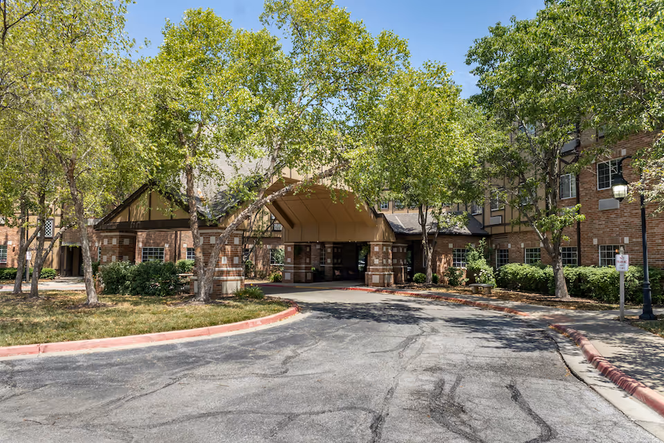Front entrance of a brick senior living building with a covered porte-cochere, trees, and a circular driveway.