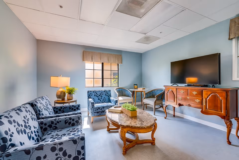A cozy senior living room with light blue walls and a window letting in natural light. The room features a patterned blue sofa and matching armchair, two additional armchairs with wooden frames, a wooden coffee table with a plant centerpiece, a side table with a lamp and plant, and a large flat-screen TV on a wooden console.