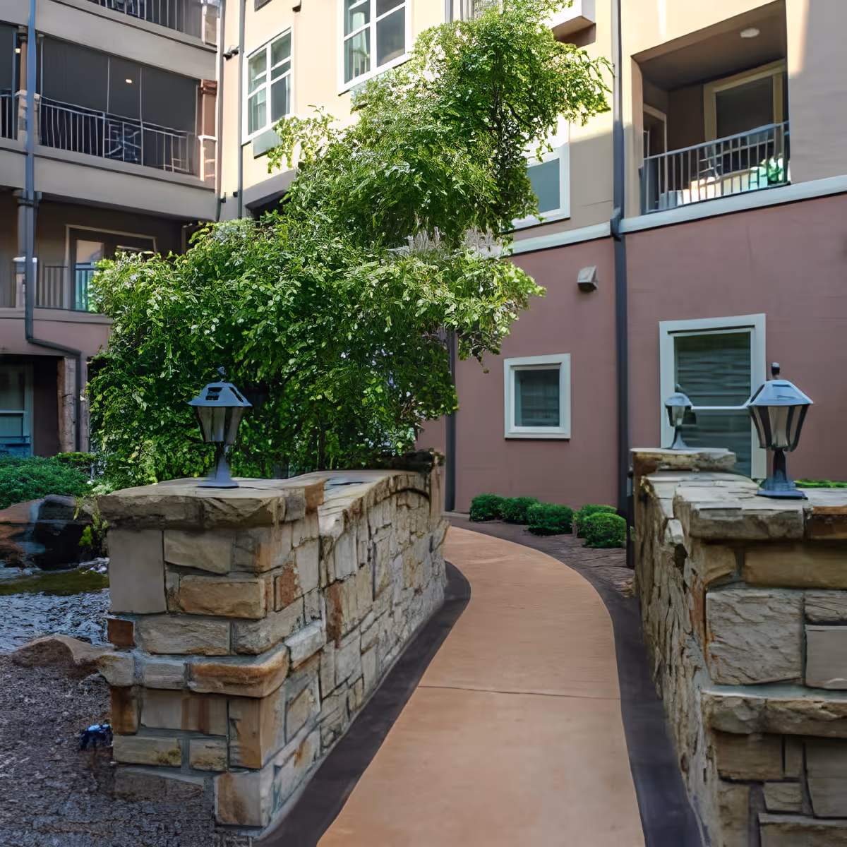A curved stone pathway with low stone walls on either side, leading through a courtyard area with green bushes and trees. The pathway is flanked by small lantern-style lights on the stone walls. Surrounding the courtyard are multi-story building walls with windows and balconies.