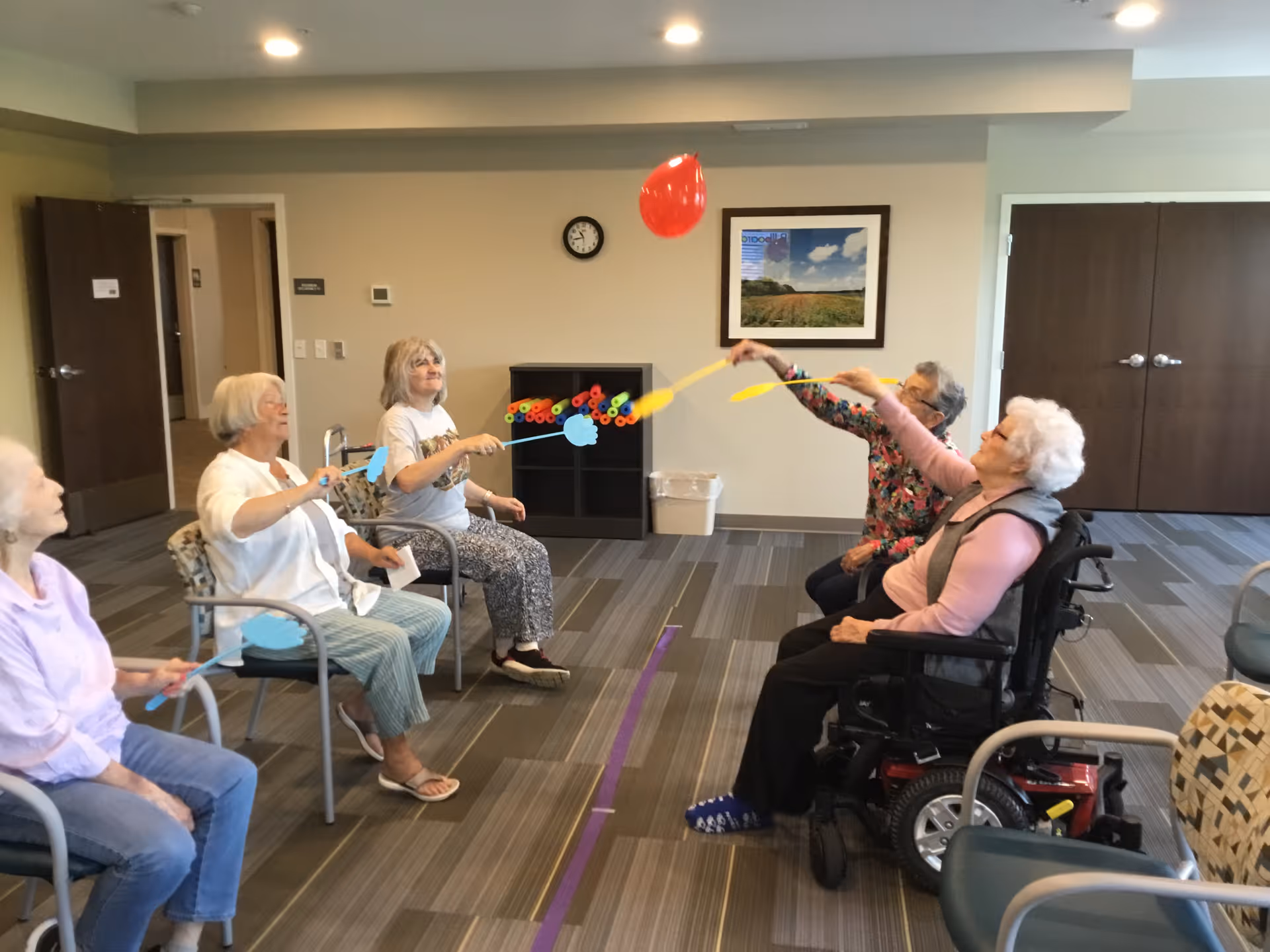 A group of elderly women seated in a circle in a common room, playing a game where they use paddles to hit a red balloon back and forth. The room has beige walls, a clock, a framed picture, and a cabinet with colorful pool noodles.