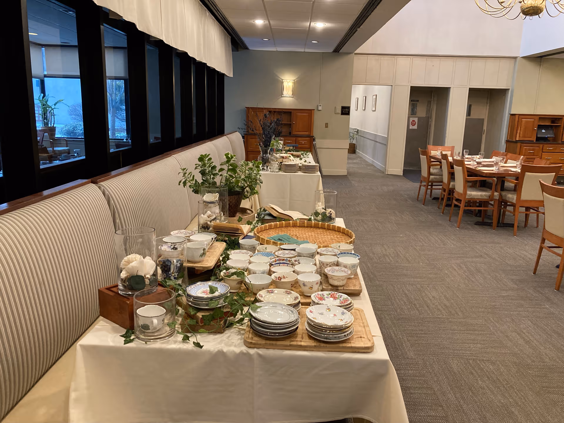 A dining area in Valley Manor Assisted Living with a long table covered in a white tablecloth displaying various floral-patterned teacups, saucers, and plates. The table is decorated with green plants and candles. In the background, there are dining tables and chairs set up, and large windows along the left side.