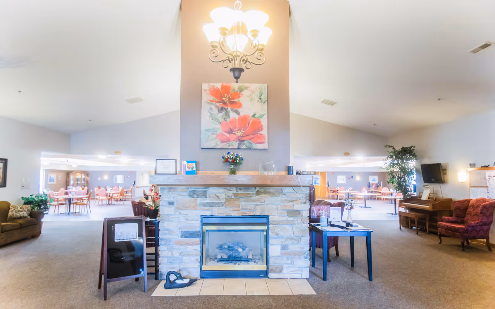 A spacious common area in an assisted living facility featuring a central stone fireplace with a floral painting and chandelier above it. Surrounding the fireplace are comfortable seating areas with armchairs and sofas, a piano, and tables. In the background, there is a dining area with multiple tables and chairs. The room is well-lit with natural and artificial light.