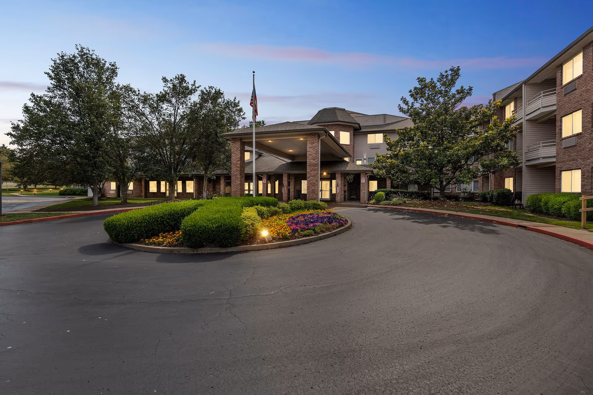 Exterior view of Holiday Uffelman Estates senior living facility at dusk, showing a circular driveway with landscaped bushes and flowers in the center, an American flag on a pole, and a brick building with multiple windows illuminated from inside.