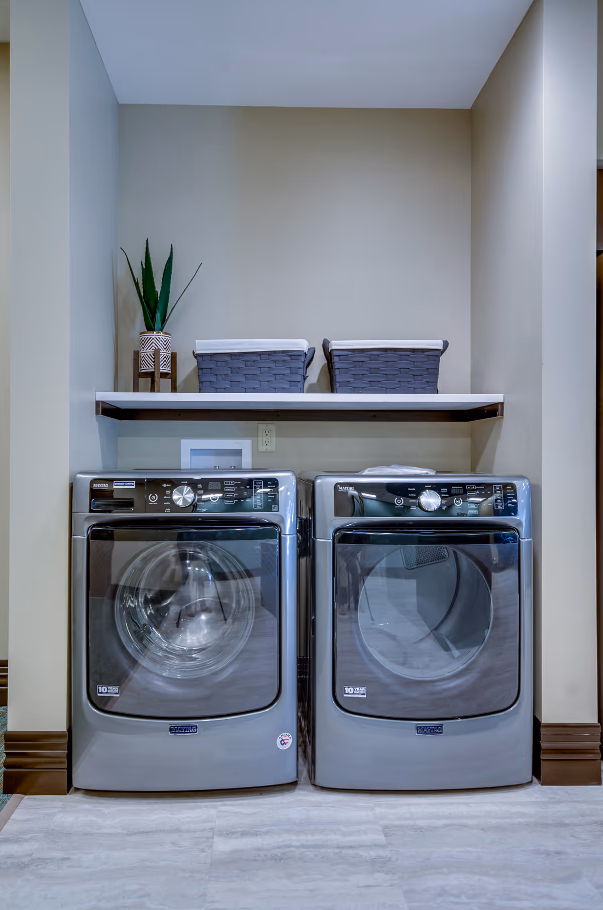 A laundry area with a front-loading washing machine and dryer side by side under a white shelf. On the shelf, there are two gray woven baskets with white liners and a small potted plant with tall green leaves. The walls are beige and the floor is light-colored tile.