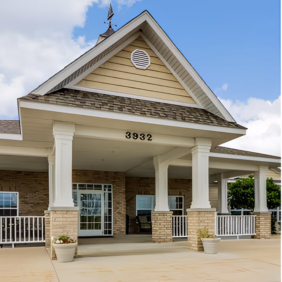 Covered entrance and porch of a senior living facility with white columns, brick pillars, the number "3932" above the portico, and potted plants.