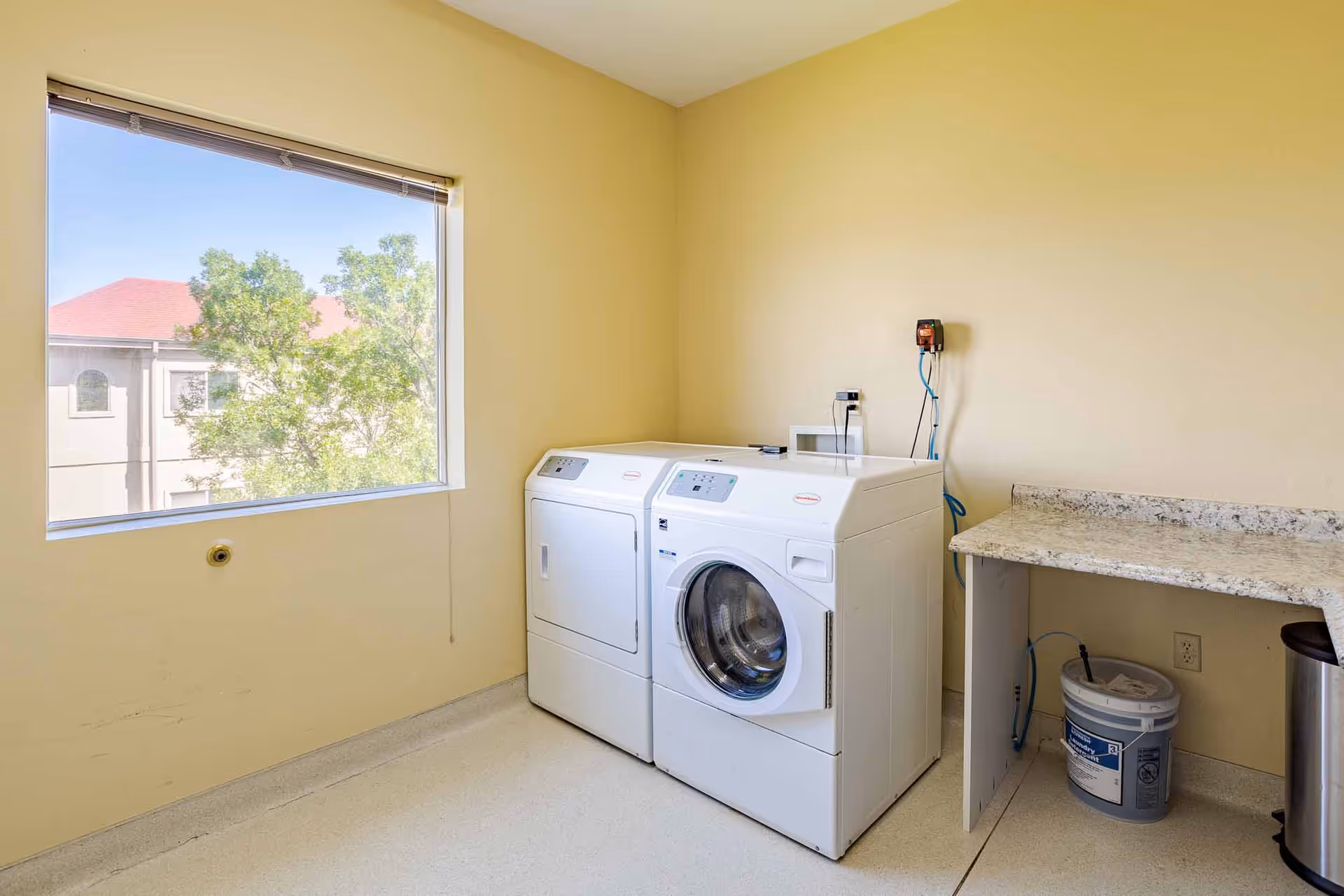 A bright laundry room with a washer and dryer beside a countertop and a large window showing trees and a neighboring building.
