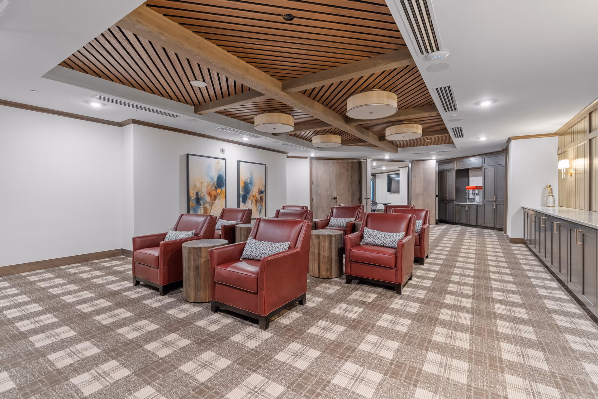 A spacious lounge area with several red leather armchairs arranged in two rows, each accompanied by a small round wooden side table. The room features a patterned carpet, wooden ceiling beams with modern circular light fixtures, and abstract artwork on the wall. There is cabinetry along one side of the room and a popcorn machine in the background.