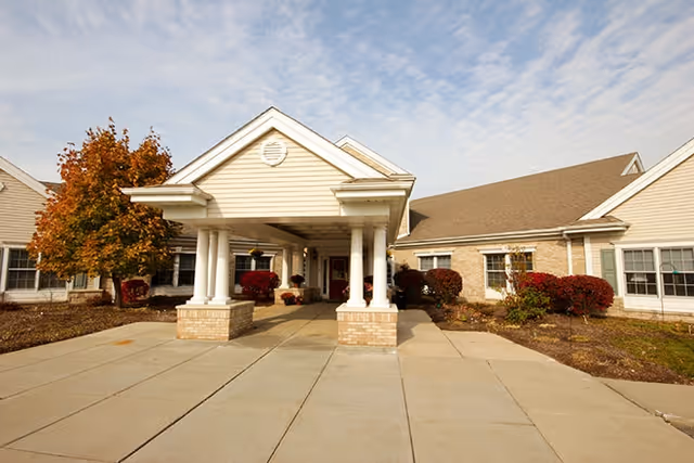 Front exterior view of a single-story senior living facility building with a covered entrance supported by white columns, surrounded by landscaped bushes and a tree with autumn foliage under a partly cloudy sky.