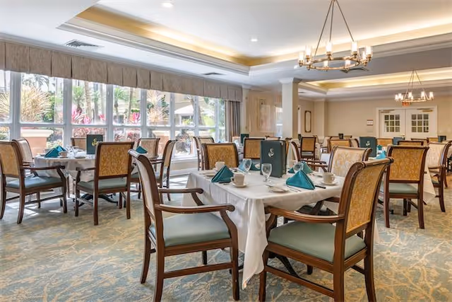 A bright dining room with multiple tables covered in white tablecloths, each set with teal napkins, cups, glasses, and silverware. The room features large windows letting in natural light, beige and green upholstered chairs, chandeliers hanging from the ceiling, and a patterned carpet.