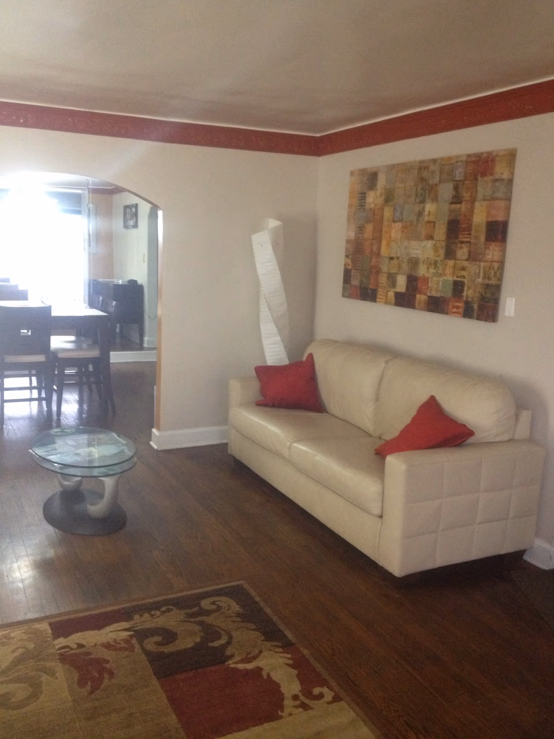 A living room with a beige leather sofa adorned with two red pillows, a glass coffee table with a unique base, a patterned area rug on wooden flooring, and a colorful abstract painting on the wall. In the background, there is an archway leading to a dining area with a table and chairs.