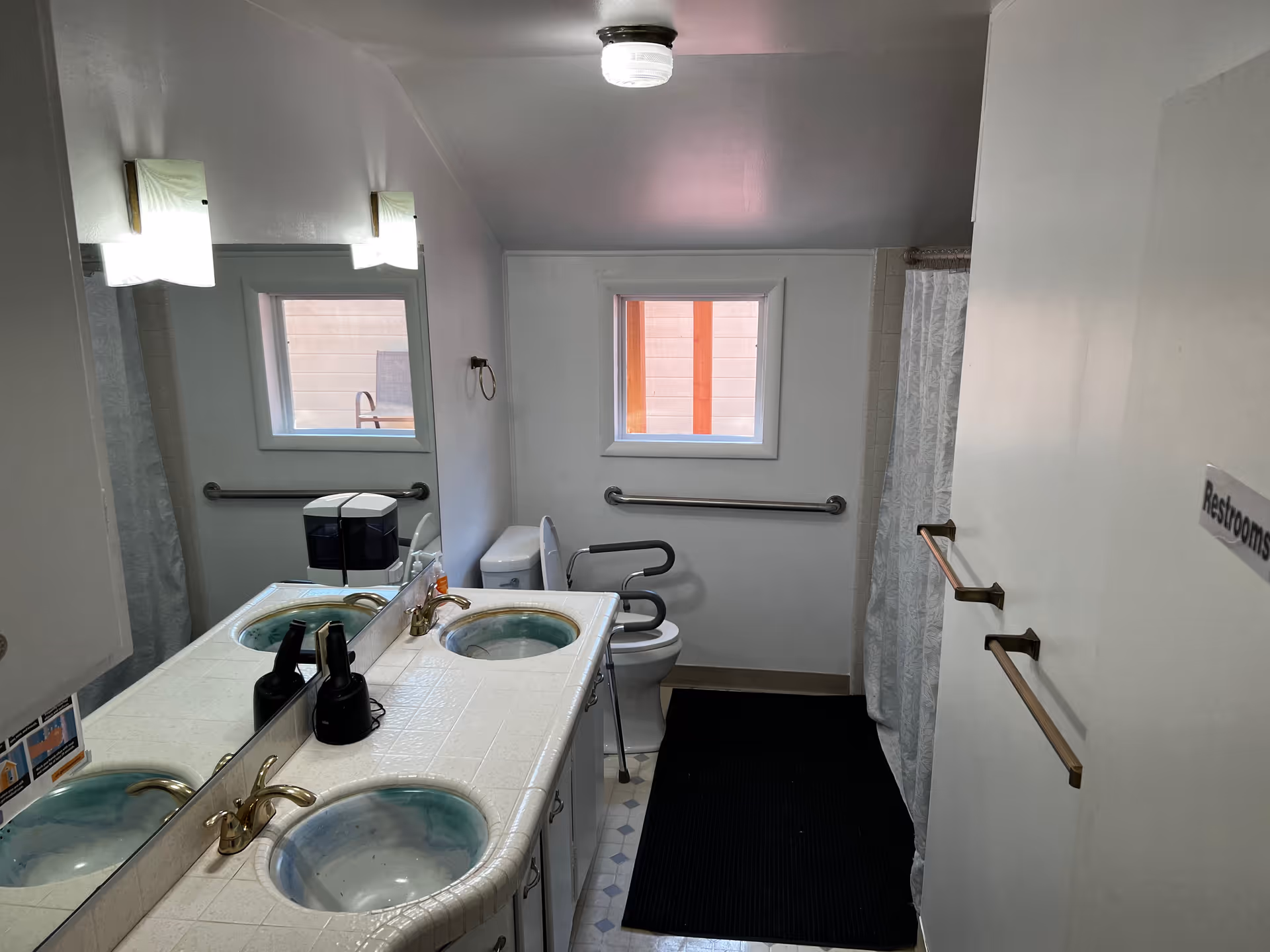 Interior view of a bathroom with two sinks on a tiled countertop, a large mirror above the sinks, a toilet with support rails, a window, and a shower area with a curtain. The bathroom has white walls and a ceiling light fixture.