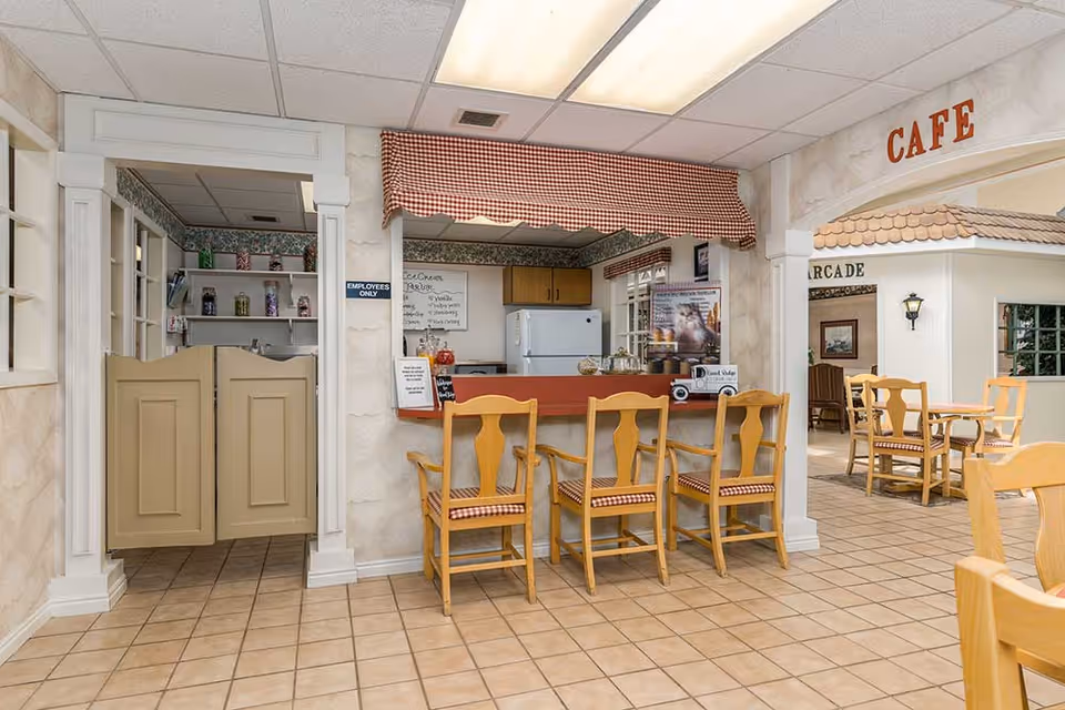 Interior view of a cozy cafe area in an assisted living facility with a counter and four wooden chairs with checkered cushions. Behind the counter is a small kitchen area with a refrigerator and shelves. To the left, there is a doorway with swinging doors labeled 'Employees Only'. To the right, there are additional tables and chairs under an archway labeled 'CAFE'.