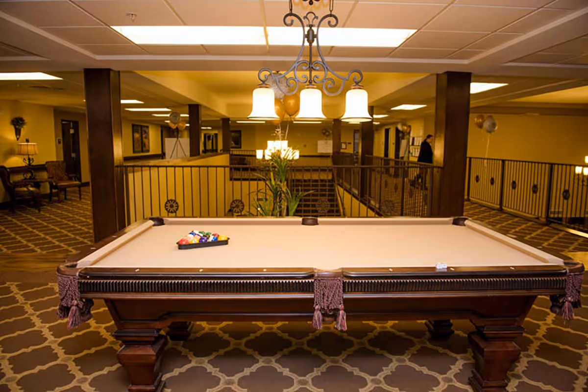 A pool table centered in a carpeted, well-lit communal lounge area with seating and railings.