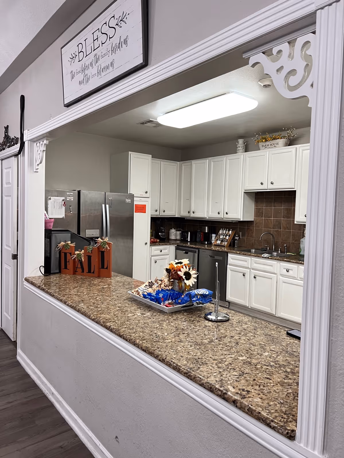 View through a pass-through window into a kitchen with white cabinets, a stainless steel refrigerator, and a granite countertop. The countertop has a decorative fall-themed block sign, a tray with snacks, and a small flower arrangement. Above the window is a sign that reads 'BLESS the seat where the family gathers and the love never ends.'